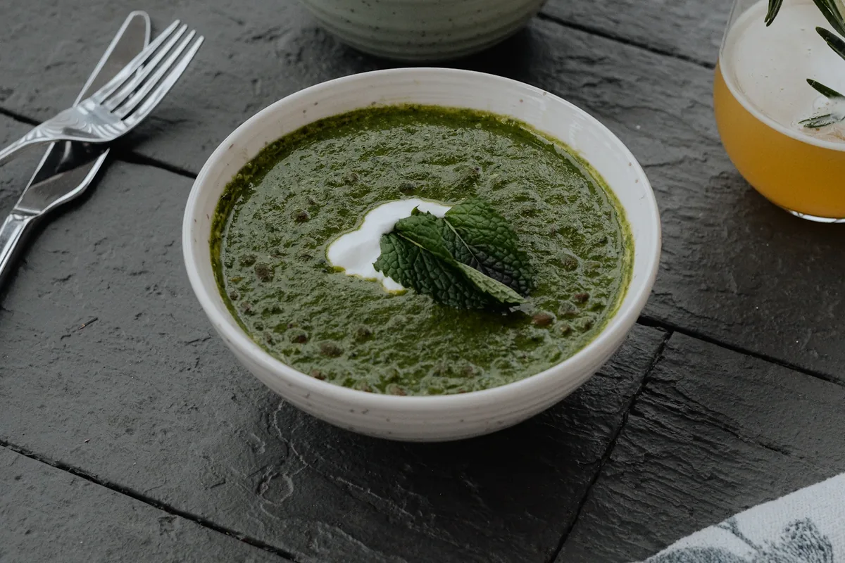 Bowl of green soup with a dollop of cream, rice and a yellow drink on a wooden table; photographer caption labels the dish lentil soup.