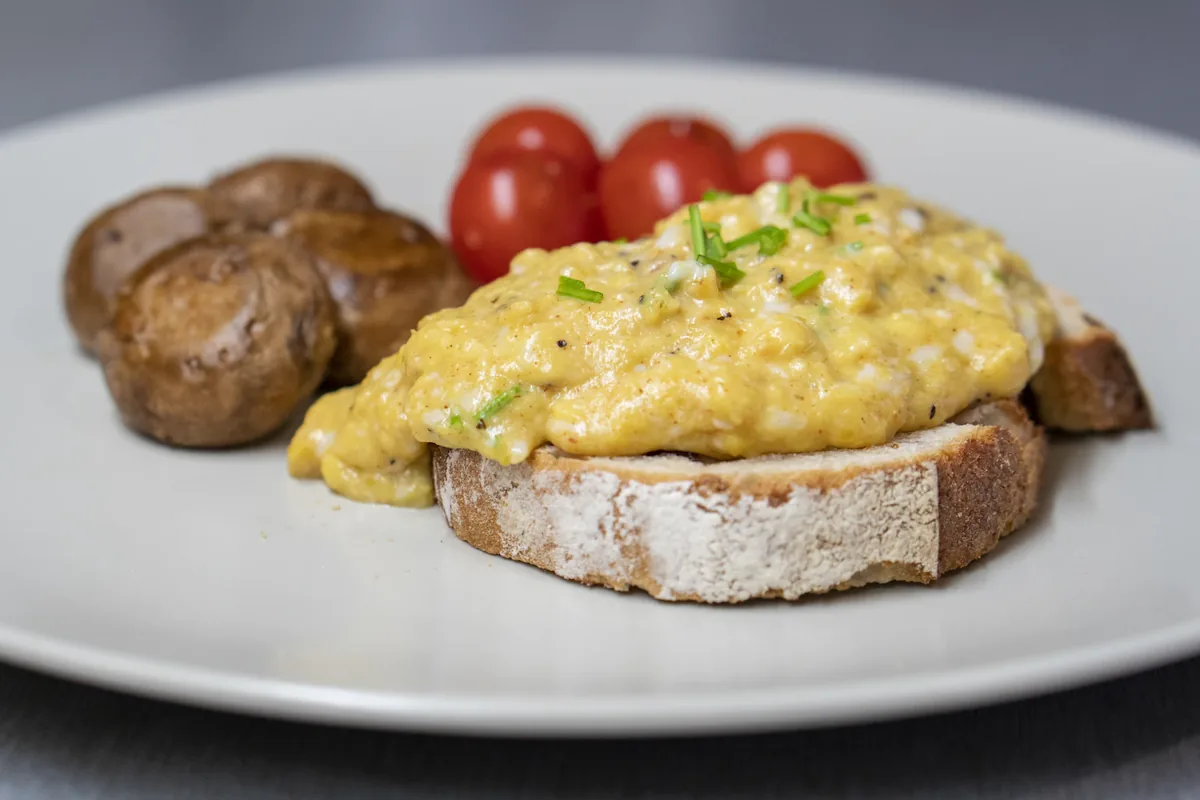 Scrambled eggs on sourdough toast with mushrooms and cherry tomatoes on a white plate; no sausage is pictured.