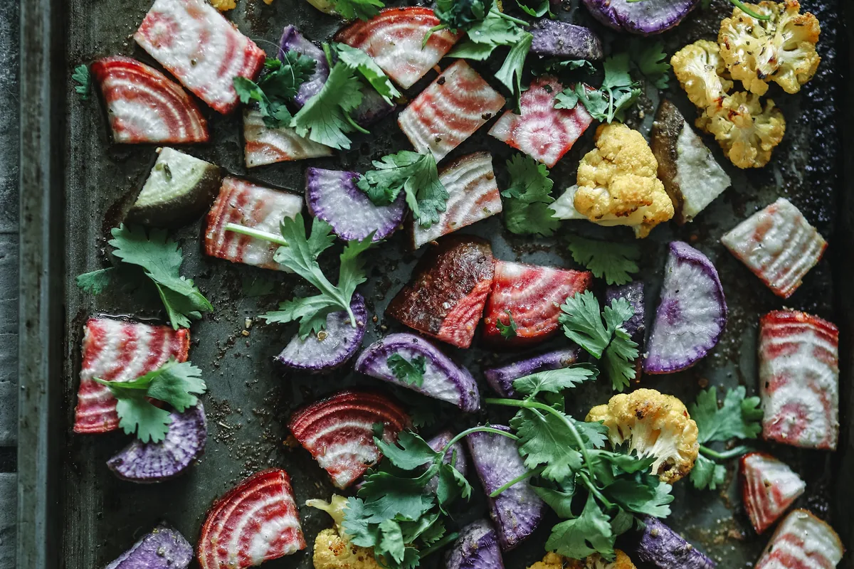 Tray of colorful roasted root vegetables and broccoli with fresh green herbs; striped pink root-vegetable slices and purple wedges are prominent.